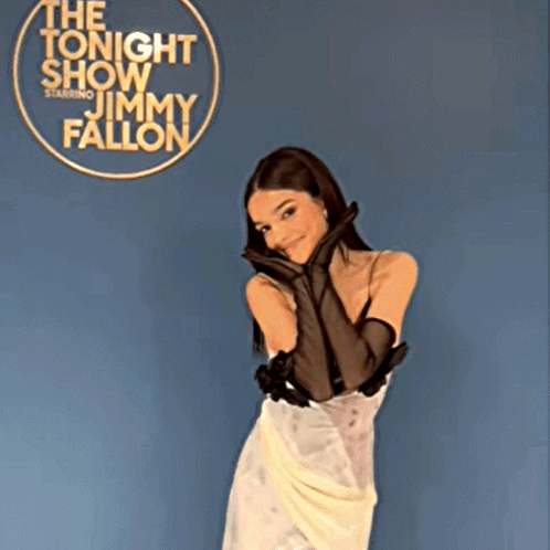 A Woman In A White Dress And Black Gloves Stands In Front Of A Sign For Jimmy Fallon GIF