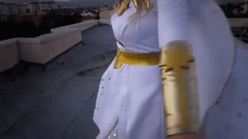 A Woman In A White Dress And Gold Gloves Is Walking On A Rooftop . GIF
