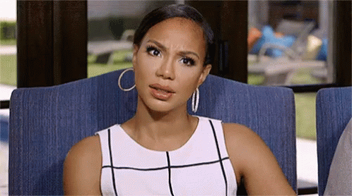 A Woman In A White Dress And Hoop Earrings Is Sitting In A Chair And Making A Funny Face . GIF