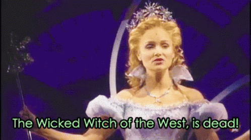 A Woman In A White Dress And Tiara Is Holding A Wand And Says The Wicked Witch Of The West Is Dead . GIF