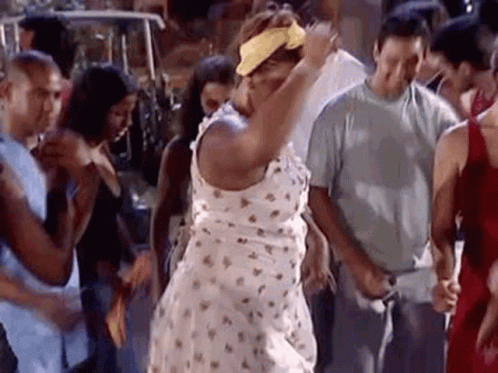 A Woman In A White Dress And Yellow Headband Is Dancing In A Crowd Of People . GIF