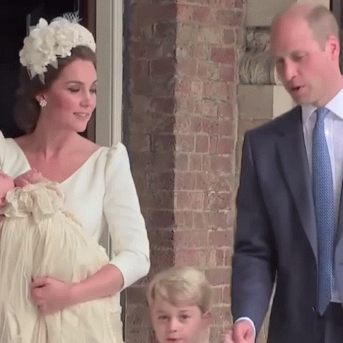 A Woman In A White Dress Holds A Baby While A Man In A Suit Looks On GIF