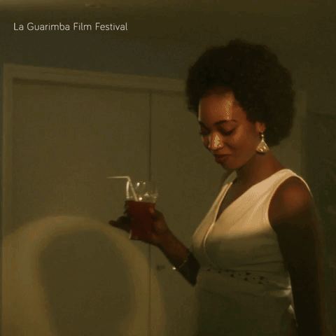 A Woman In A White Dress Holds A Glass In Front Of A La Guarimba Film Festival Poster GIF