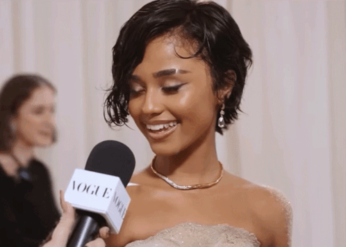 A Woman In A White Dress Is Being Interviewed By A Vogue Microphone GIF