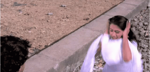 A Woman In A White Dress Is Covering Her Ears With Her Hands While A Man Looks On . GIF