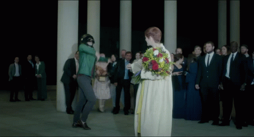 A Woman In A White Dress Is Holding A Bouquet Of Flowers In Front Of A Crowd Of People GIF