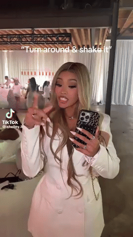 A Woman In A White Dress Is Holding A Cell Phone In Her Hand And Says " Turn Around & Shake It " GIF