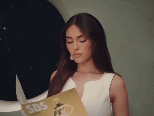 A Woman In A White Dress Is Holding A Magazine That Says Sbs GIF