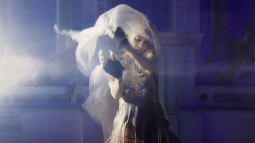 A Woman In A White Dress Is Hugging Another Woman In A Dark Room GIF