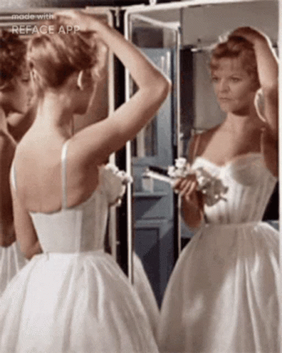 A Woman In A White Dress Is Looking At Her Reflection In A Mirror GIF