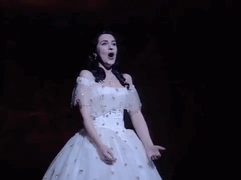 A Woman In A White Dress Is Singing On A Stage With Her Mouth Open . GIF