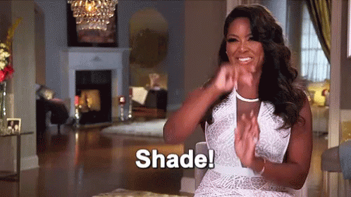A Woman In A White Dress Is Sitting In A Living Room Clapping Her Hands And Says Shade . GIF