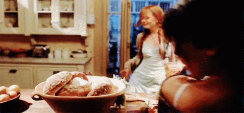 a woman in a white dress is standing in a kitchen with a bowl of bread on the table .