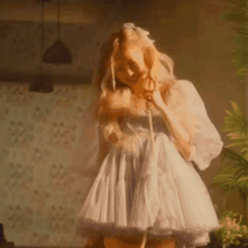 A Woman In A White Dress Is Standing In A Room Talking On A Telephone . GIF