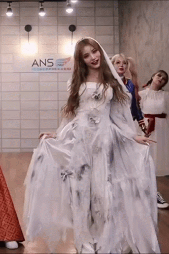 A Woman In A White Dress Is Standing In A Room With Other Women Dressed In Costumes . GIF