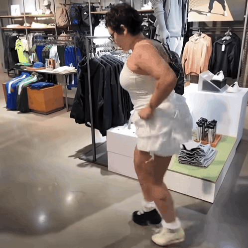 A Woman In A White Dress Is Standing In Front Of A Clothing Rack GIF