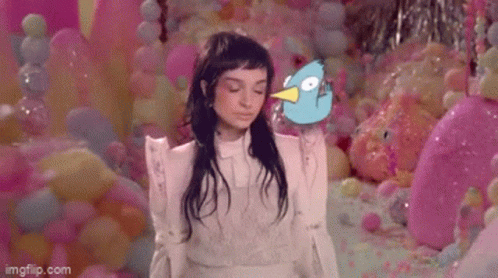 A Woman In A White Dress Is Standing In Front Of A Wall Of Pink Balloons Holding A Blue Bird . GIF