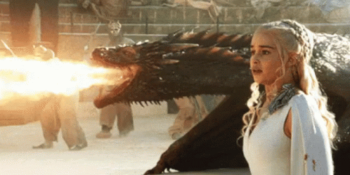 A Woman In A White Dress Is Standing Next To A Dragon That Is Shooting Fire From Its Mouth . GIF