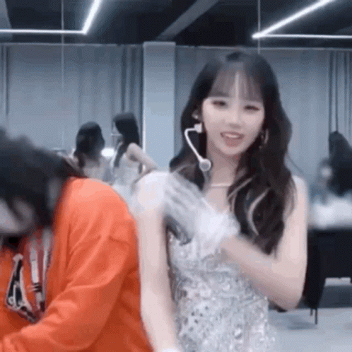 A Woman In A White Dress Is Standing Next To A Man In An Orange Sweatshirt In A Room . GIF