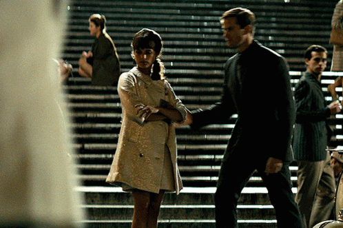 A Woman In A White Dress Is Standing Next To A Man On A Set Of Stairs GIF