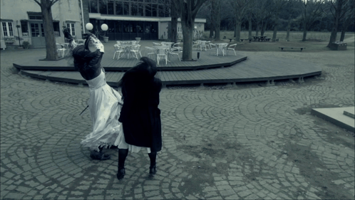 A Woman In A White Dress Is Standing On A Cobblestone Walkway GIF