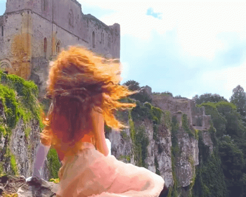 A Woman In A White Dress Is Standing On A Rocky Cliff With A Castle In The Background GIF