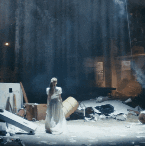 A Woman In A White Dress Is Standing On A Stage With A Barrel In The Background GIF