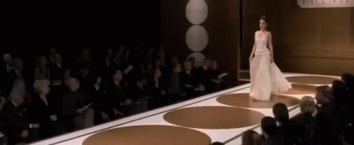 A Woman In A White Dress Is Walking Down A Runway In Front Of A Crowd Of People . GIF