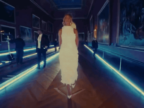 A Woman In A White Dress Is Walking Through A Dark Hallway GIF