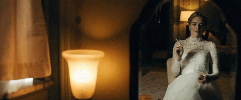 A Woman In A White Dress Smoking A Cigarette In Front Of A Mirror GIF