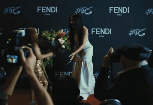 A Woman In A White Dress Stands In Front Of A Wall That Says Fendi Roma GIF