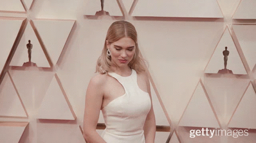 A Woman In A White Dress Stands In Front Of A Wall With Oscar Statues GIF
