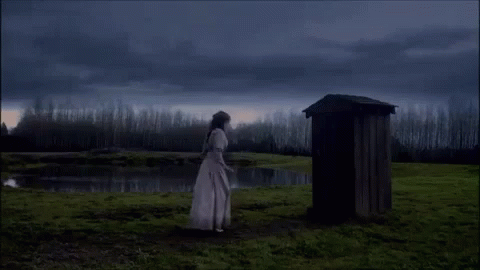 A Woman In A White Dress Stands In Front Of A Wooden Outhouse In A Field GIF