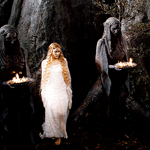 A Woman In A White Dress Stands In Front Of Two Statues Holding Candles GIF