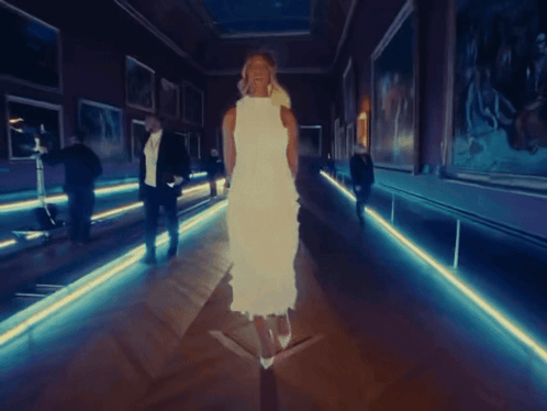 A Woman In A White Dress Walks Down A Hallway GIF