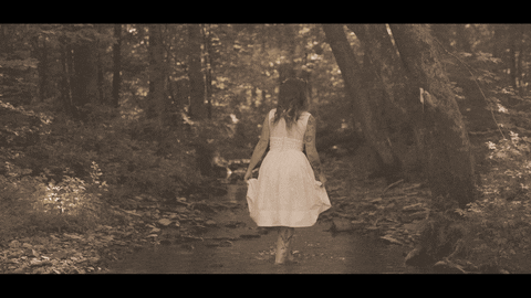 A Woman In A White Dress Walks Through The Woods GIF
