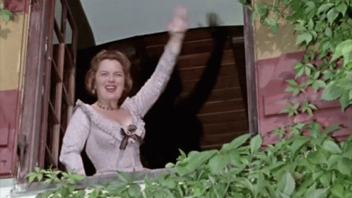 A Woman In A White Dress Waving From A Balcony GIF
