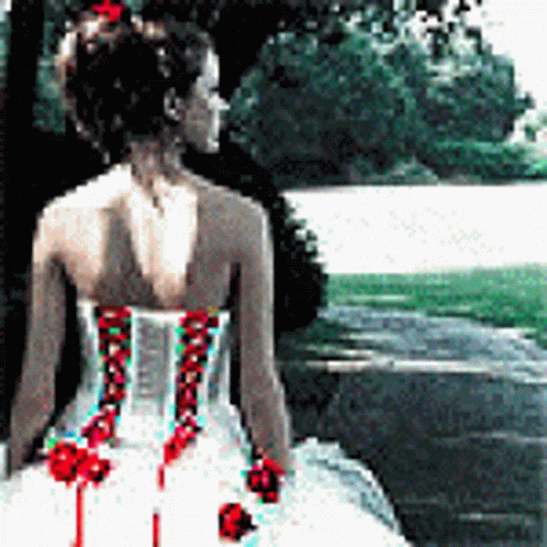 A Woman In A White Dress With Red Flowers On The Back Is Standing In Front Of A Lake GIF