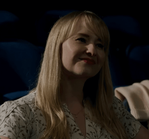 A Woman In A White Floral Shirt Is Sitting In A Dark Room GIF
