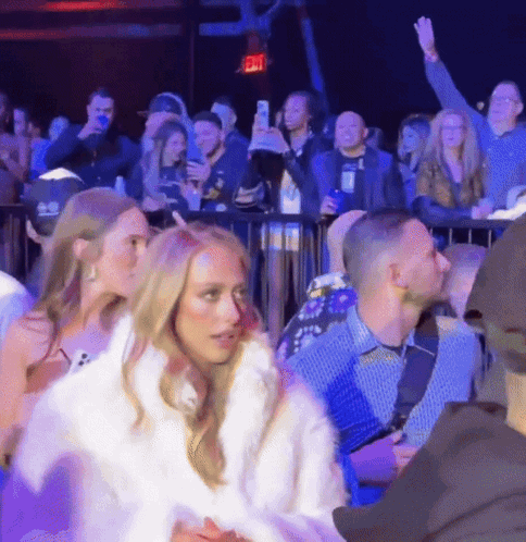 A Woman In A White Fur Coat Stands In Front Of A Crowd With An Exit Sign Above Her Head GIF