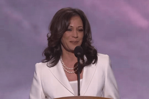 A Woman In A White Jacket And Pearl Necklace Stands At A Podium Speaking Into A Microphone GIF
