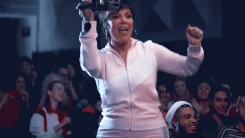 A Woman In A White Jacket Is Standing In Front Of A Crowd With Her Fist In The Air GIF