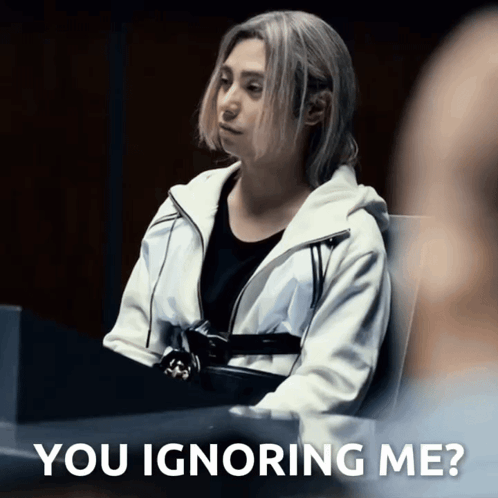 A Woman In A White Jacket Sits At A Table With The Words " You Ignoring Me " On The Bottom GIF