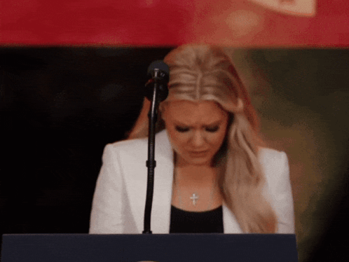 A Woman In A White Jacket Stands At A Podium With Her Eyes Closed GIF