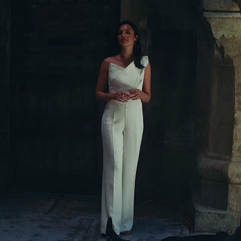A Woman In A White Jumpsuit Is Standing In Front Of A Door GIF