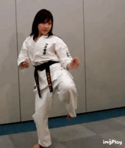 A Woman In A White Karate Uniform With A Black Belt Is Kicking A Ball . GIF