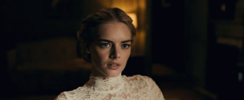 A Woman In A White Lace Dress Looks At The Camera In A Dark Room GIF