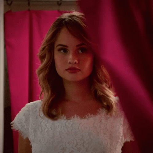 A Woman In A White Lace Top Is Looking Through A Pink Curtain GIF