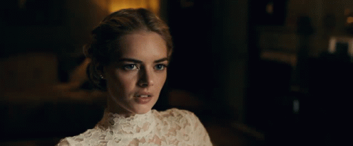 A Woman In A White Lace Top Looks At The Camera In A Dark Room GIF