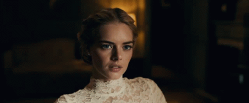 A Woman In A White Lace Top Looks At The Camera In A Dark Room GIF